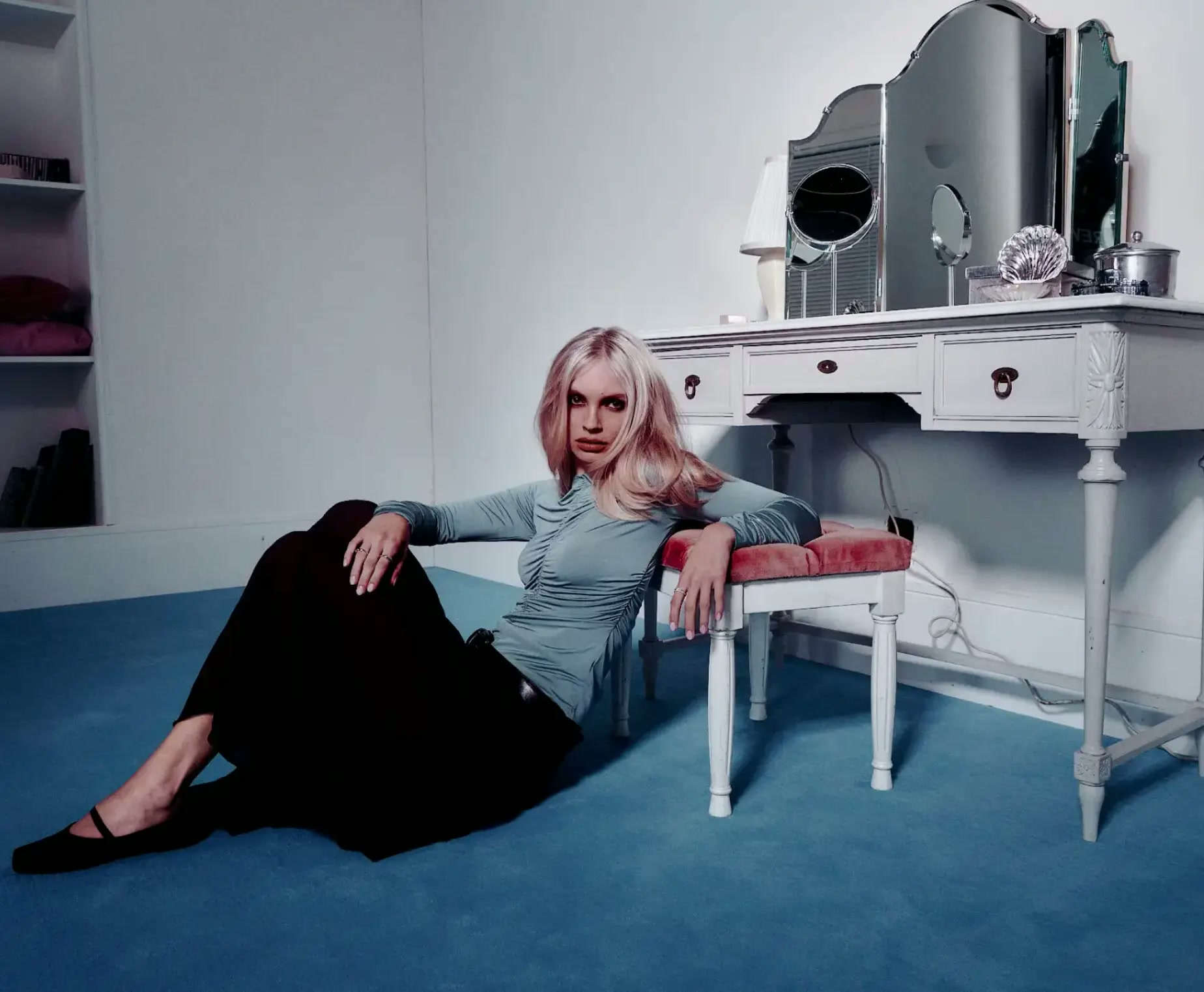 Model seated on a blue-carpeted floor beside a white dressing table